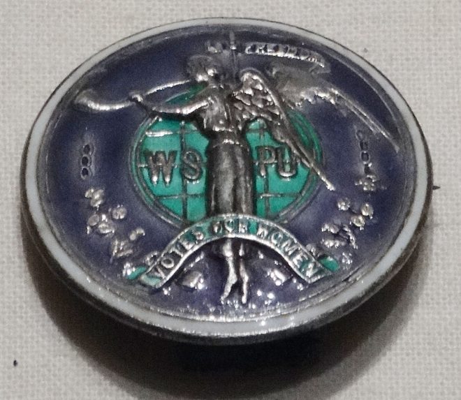 Suffragette badge in green, white and purple, the colours most associated with the Women's Social and Political Union., one of the main Suffragette organisations under the leadership of Emmeline Pankhurst. On display in Scotland a Changing National at National Museum of Scotland.