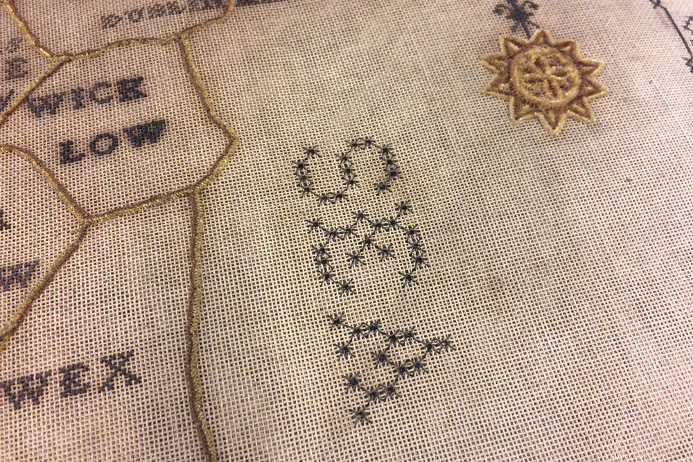 A detail of the map showing the use of cross stitch to create text in different sizes and fonts whilst the borders and compass are of laid chenille threads which has a more 3D, textural finish.