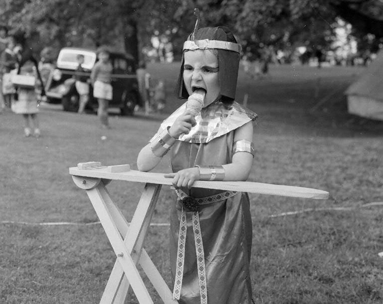 A small child in a field at a Gala Day dressed in Egyptian costume licks ice cream on a cone. 
