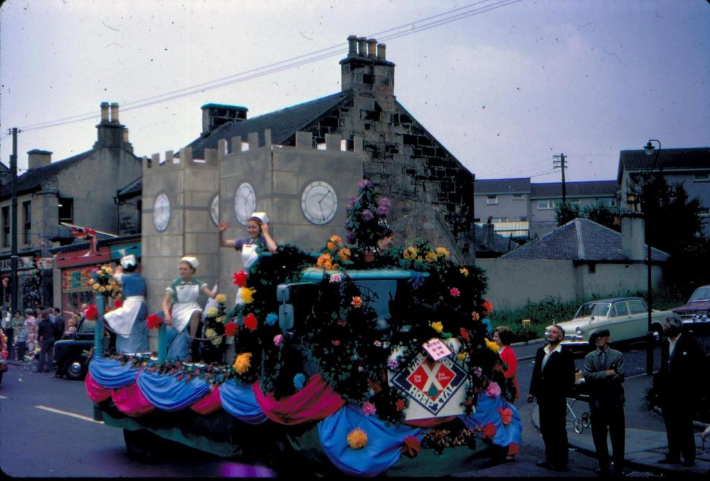 Grainy photograph of a parade float with women in nurse outfits going down a village road on a Gala Day.