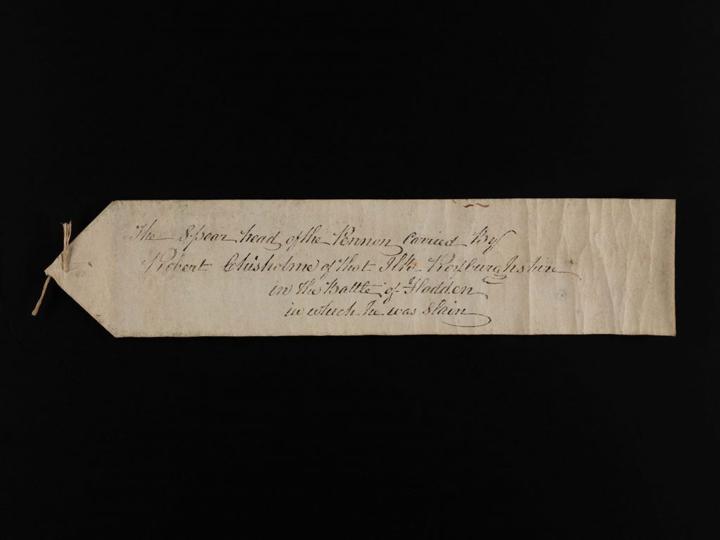 Off-white rectangular label with one triangular end with old fashioned cursive writing describing the Flodden spearhead.