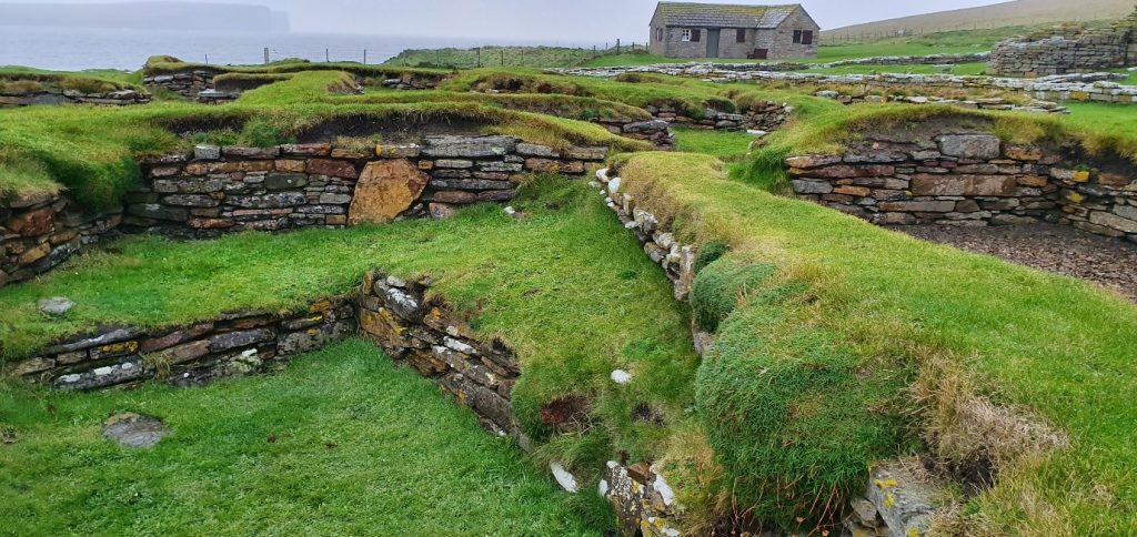 Ruinous Viking settlement at Brough of Birsay. Stone-lined pits mark where buildings used to be. Orkney sea and cliffs are in the distance.