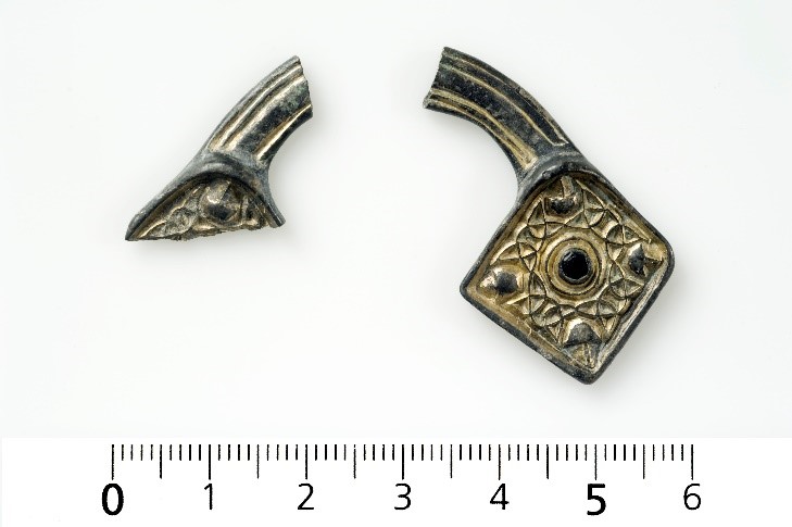 Two small fragments of a dark metal brooch, its ends decorated with vine-like patterns.