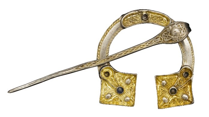 Golden brooch with a long, sharp pin angled diagonally across a horseshoe-shaped brooch with an opening at the bottom and pearl-like jewels.