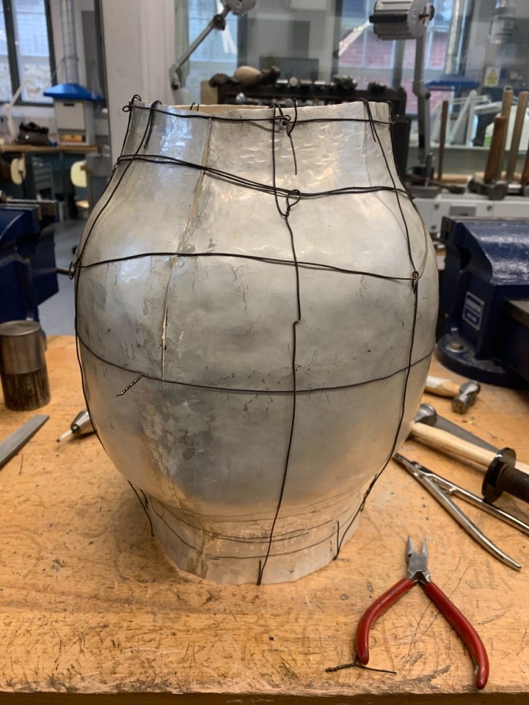 The now bulbous 'cylinder' standing on a work table, surrounded by tools including plyers and hammers. The sculpture is tightly wound with black wire. 