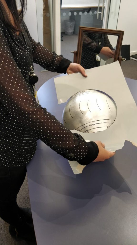 The artist holds a piece of card that has a semi circle cut our of it over the sculpture. The silver sculpture has markings on it in black pen. 