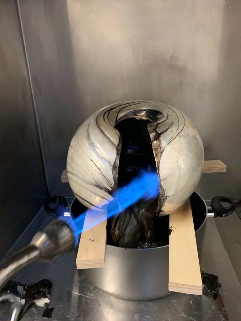 The 'C' shaped sculpture is resting on some wooden planks with s large kitchen pot beneath it. A blow torch is being applied to the black 'tar' substance in the middle of the sculpture, which is melting into the pan below. 