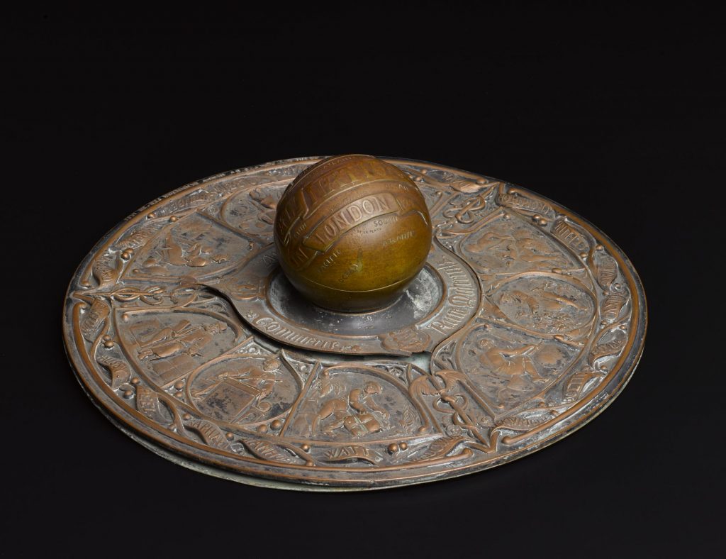 A round flat brass and copper inkwell with a spherical handle in the middle. The 'flat' section is decorated with scenes that include a potter and a weaver at work, with names of artists, inventors and designers in a ribbon around the edge.