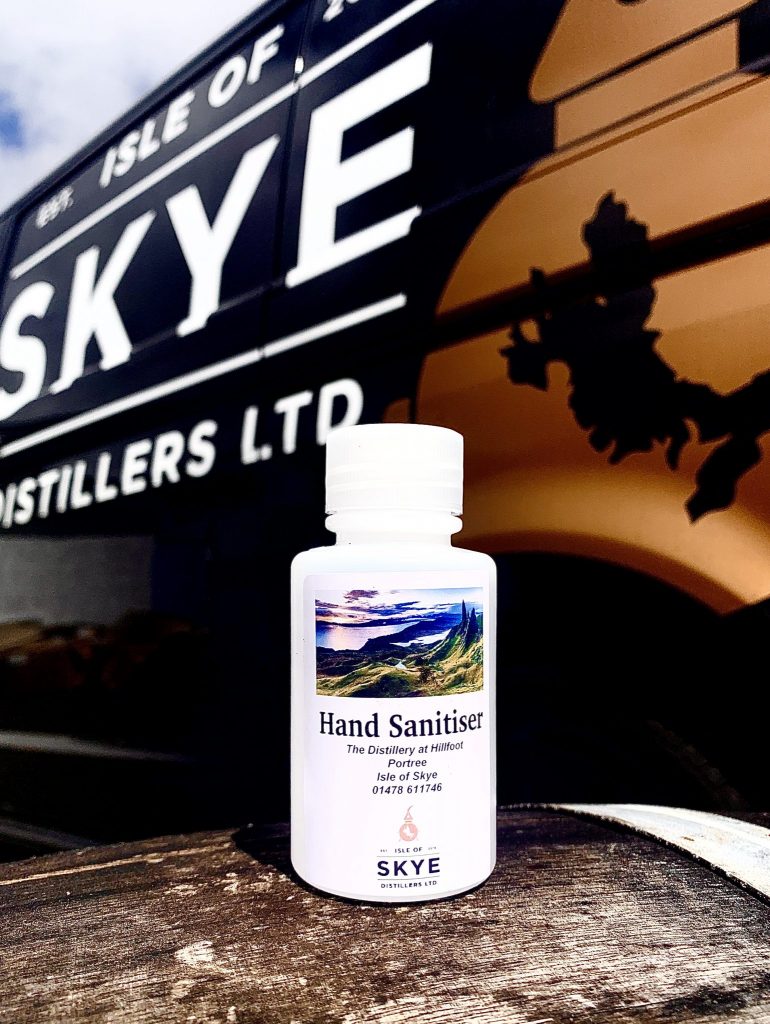 A bottle of hand sanitiser sitting on a table. The label has a photo of the Isle of Skye on it and behind the bottle is a banner for the Isle of Skye distillers.