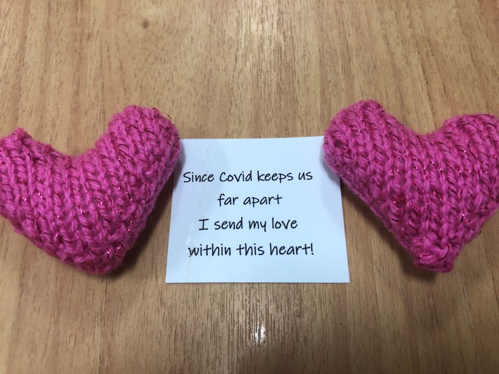 Two pink knitted hearts next to each other on a table. Between them is a piece of paper with the words "ince COVID keeps us far apart I send my love within this heart" written on it.