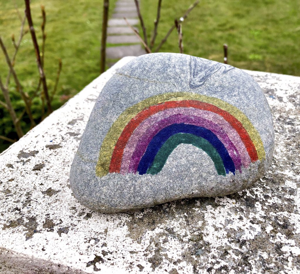 A grey pebble sitting on a stone with grass behind it. The pebble has a stylised rainbow drawn on it.