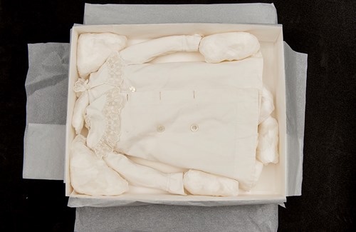 A creamy white double-breasted coat with a lacy collar laid flat in a box. It's packed with tissue paper and packaging to keep it in place so it doesn't shift about. 