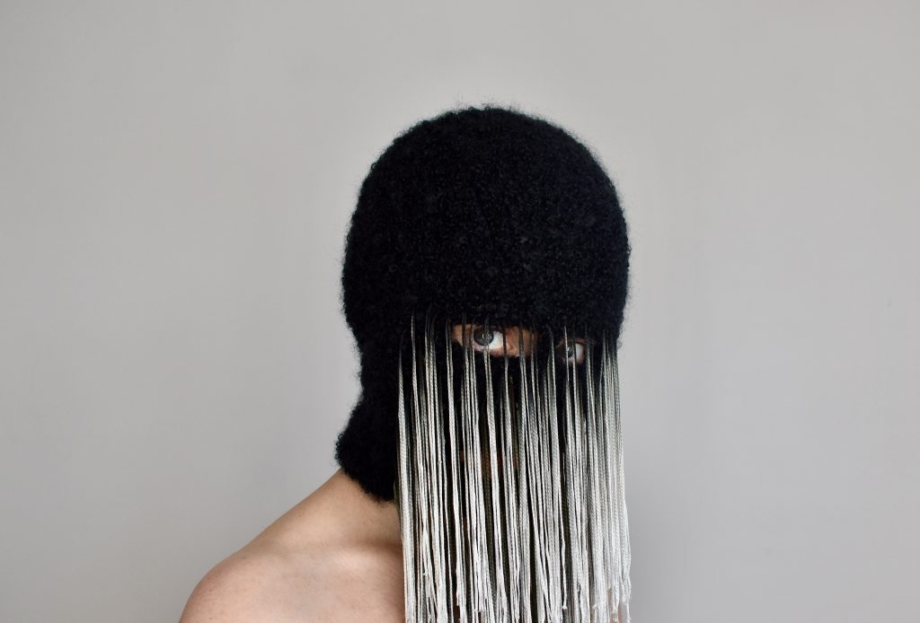 The artist wears a black balaclava-style mask with wispy white tendrils hanging in front of it in a veil-like fashion.  