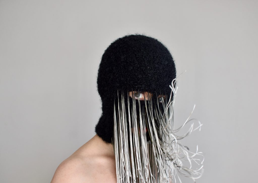 The artist wears a black balaclava-style mask with wispy white tendrils hanging in front of it in a veil-like fashion.  The tendrils are moving with her breath. 
