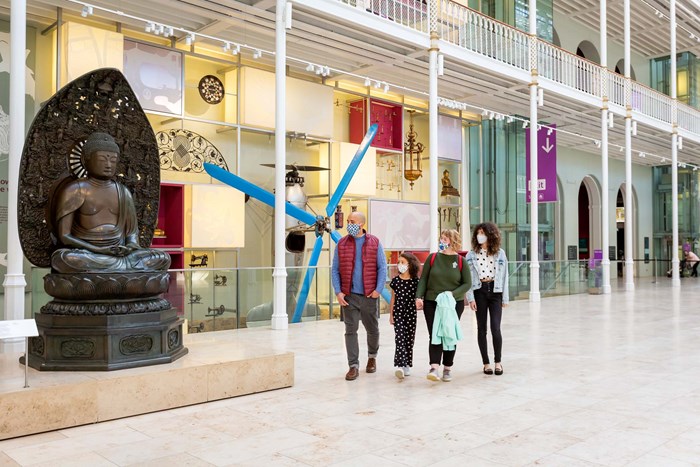 The Grand Gallery of the National Museum of Scotland showing people walking and pointing, with a large Buddha statue on display. 