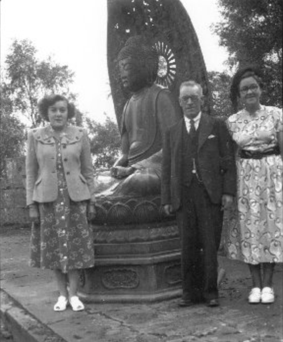 A black and white photograph showing three people standing in front of one of the Buddha statues with their backs to it. 
