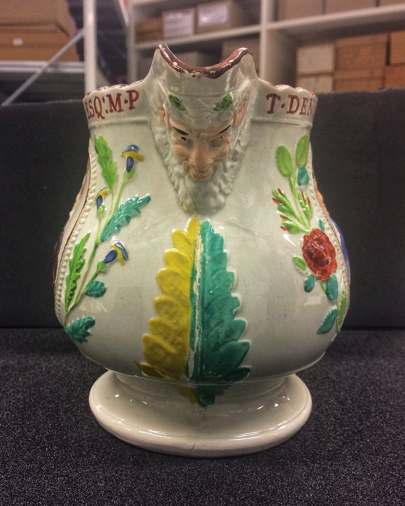 Jug, like a gravy jug, facing forward on a grey surface. Floral motifs decorate the sides and a god-like face decorates the spout like a ship's prow figurehead.