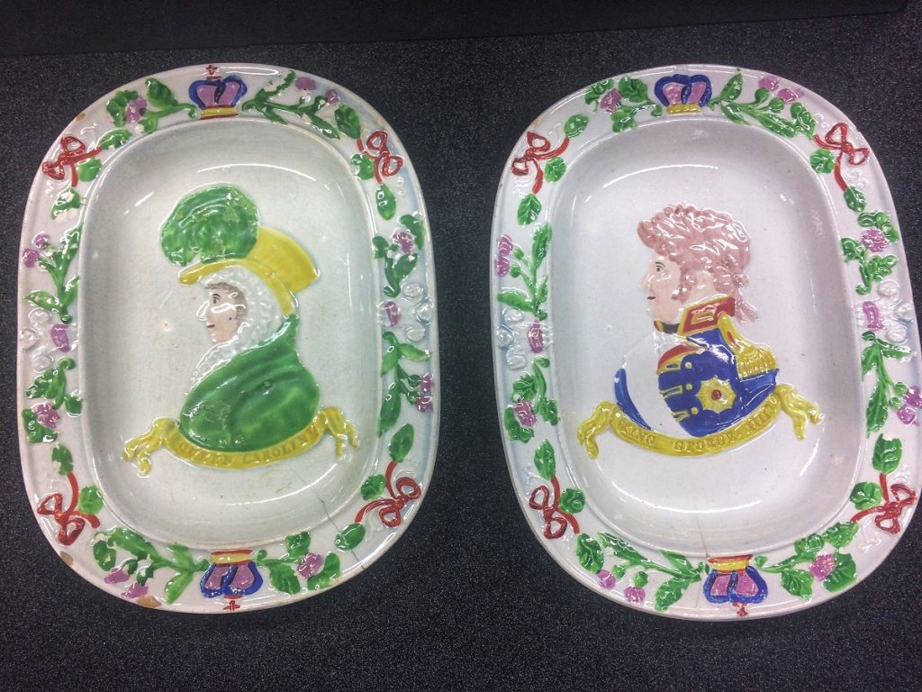 Two oval dishes side by side. Left dish depicts Caroline in green dress with yellow hat, right dish depicts George VI in blue military uniform with no hat. 