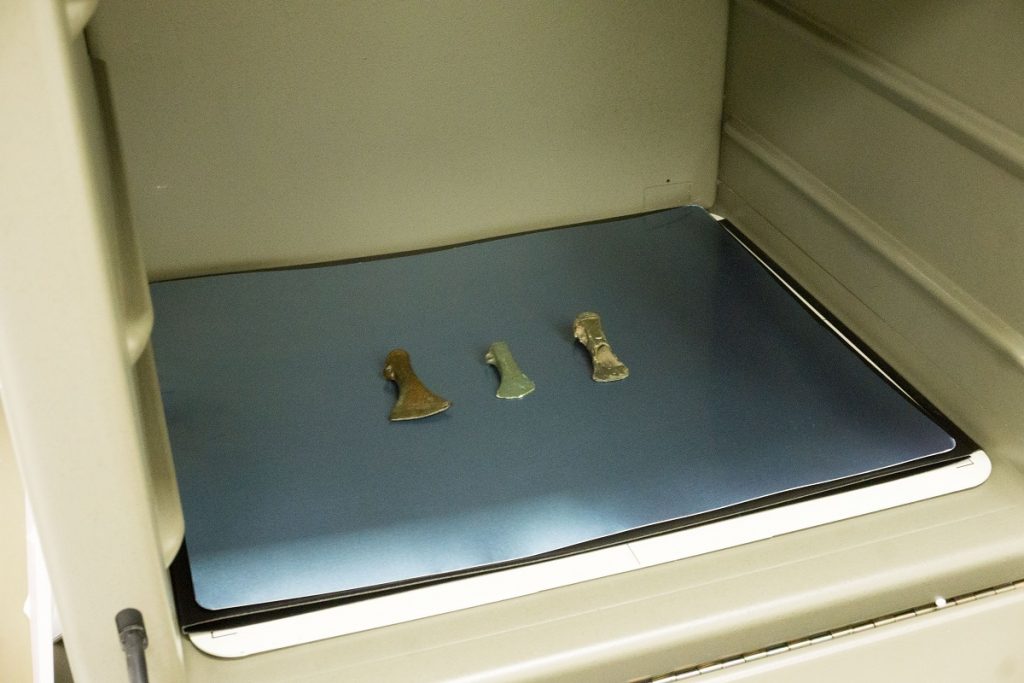 Three brown to green axeheads sit on a grey plate within an x-ray machine that looks like inside a microwave.