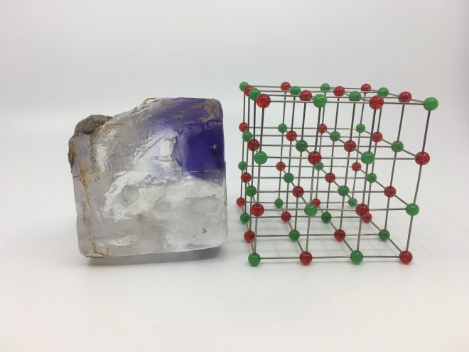 A roughly square chunk of grey and bright blue mineral sits on a white surface next to a lattice-like cube.