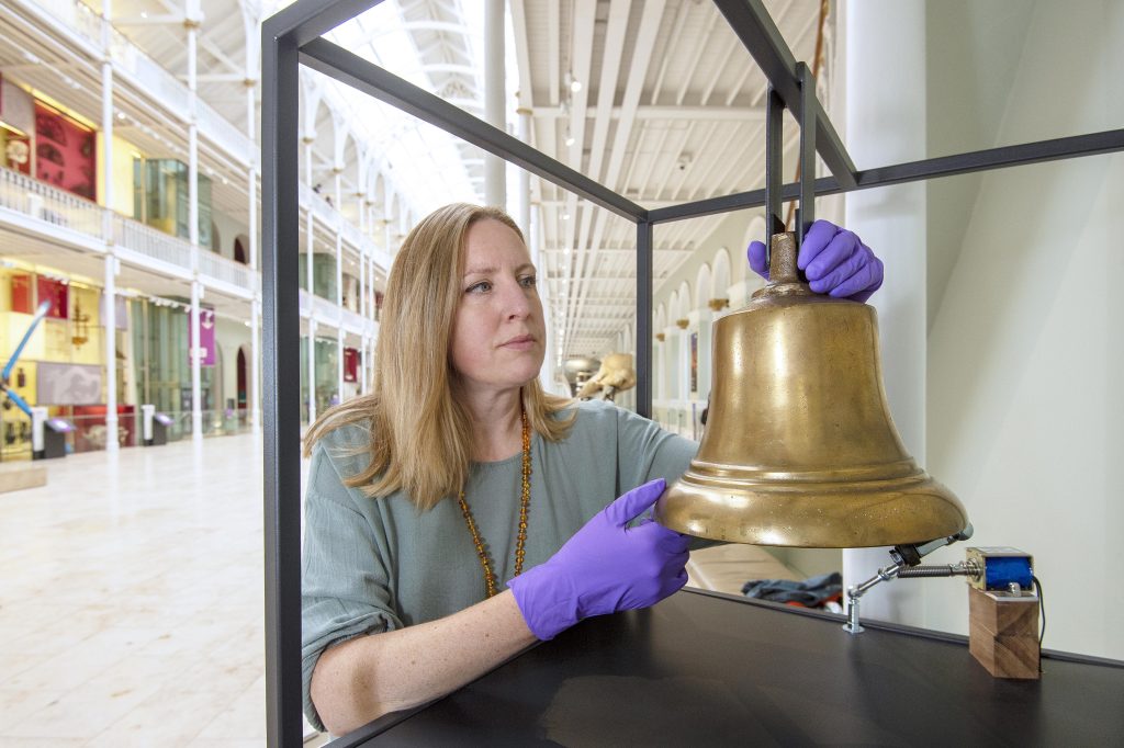 A woman stands in the Grand Gallery with a large bell and she's touching it with purple gloves on.