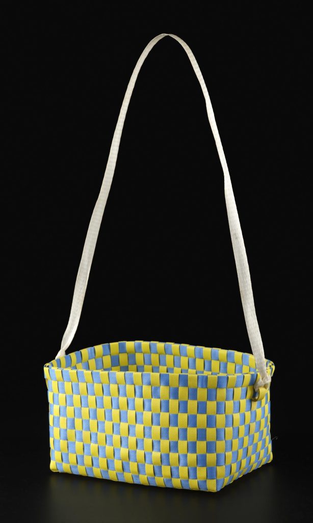 Wide and fairly shallow basket with yellow and blue checkerboard pattern and a long strap, as if hanging on a post, against a black background.