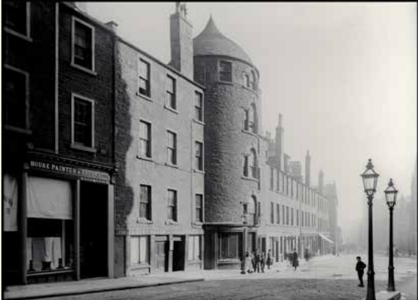 Black and white photo of a city centre street from the late 1870s with tenements and shops.