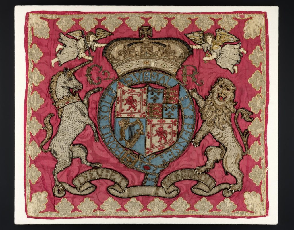 Embroidered banner, primarily red and gold. A unicorn on the left and a lion on the right hold up a coat of arms with lions, a harp and fleur-de-lis topped by a crown.
