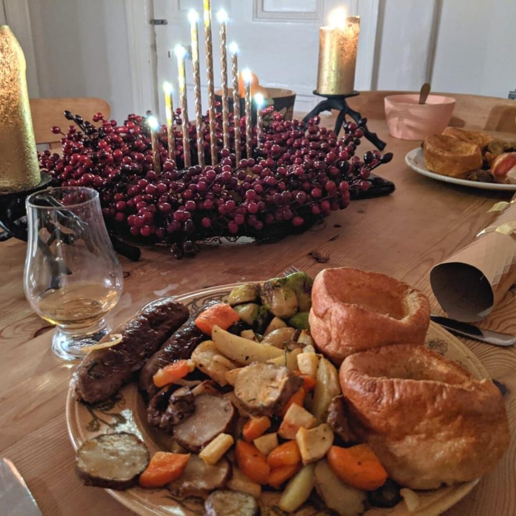 Christmas dinner table with a plate full of Yorkshire puddings, roast vegetables and sausages, with candles and a wreath in the background.