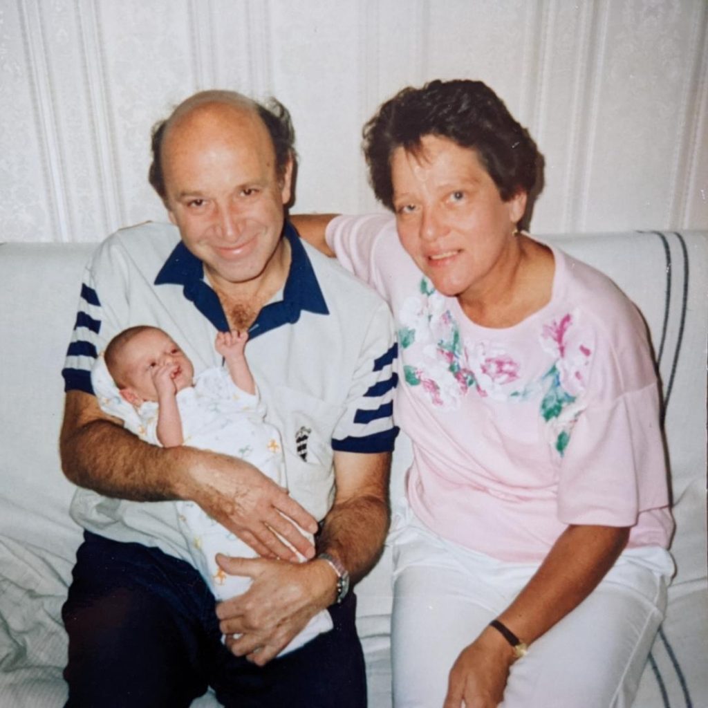 A man and a woman, middle aged, sitting on a white couch smiling while holding a newborn baby, their grandson.