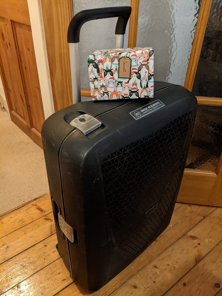 A large black suitcase standing upright in a hallway with wooden floors, topped by a present.