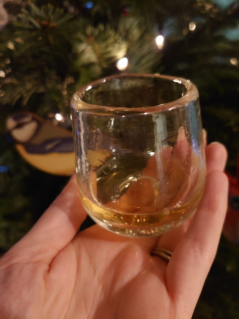 A small whisky glass is held up in font of a Christmas tree with warm lights.