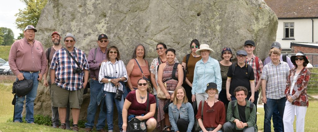 Large group of people standing in front of a huge boulder in a grassy field on a warm, sunny day.