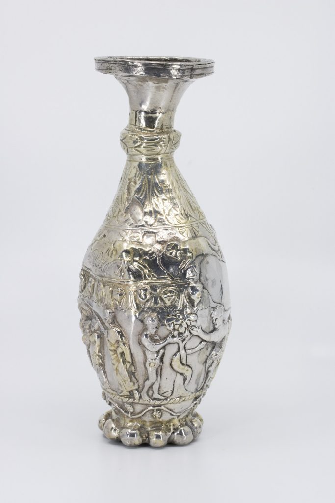 Very shiny silver flagon, shaped like a flower vase. Bulging in the middle, biblical scenes adorn it surface including Adam and Eve taking the apple.