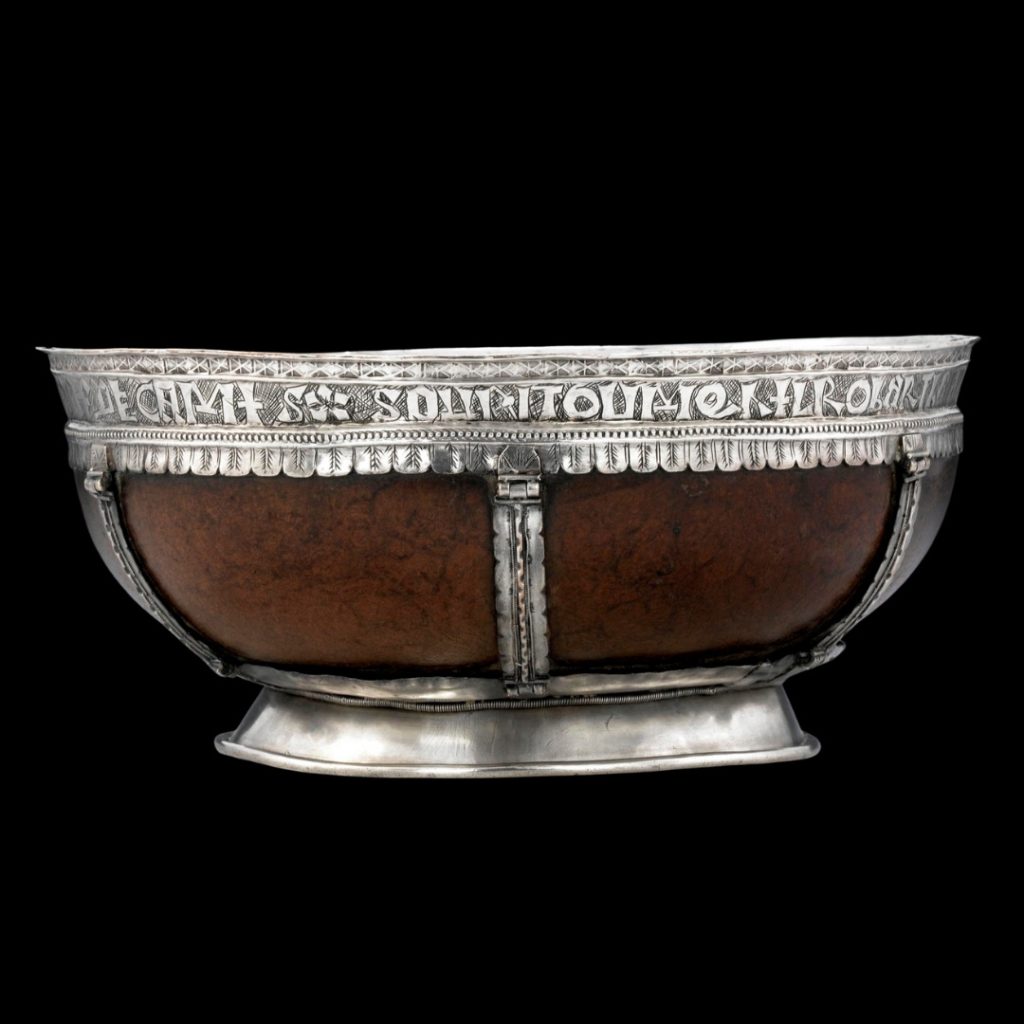 Wide bowl with a brownish-red hue contained within an enclosure of silver covered with small designs and letters across the top rim.