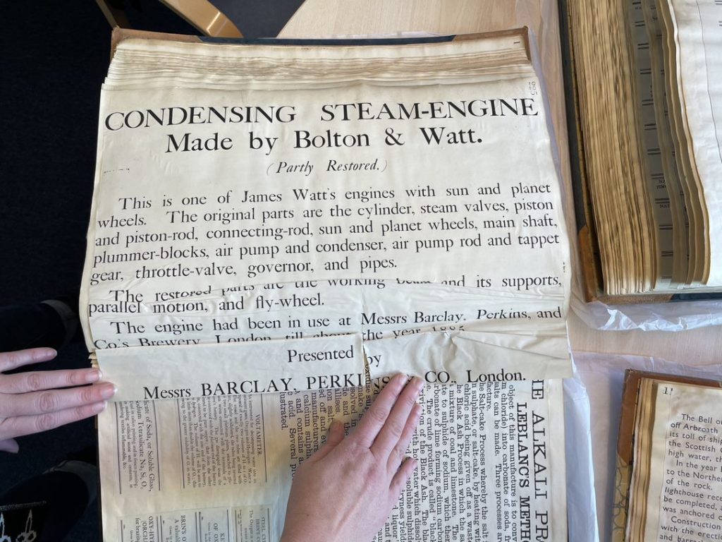 Colour photo of a large book with cream printed labels pasted inside describing the Condensing Steam-Engine made by Bolton & Watt. 