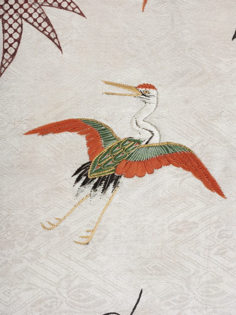 Close up of an embroidered crane in flight. Orange wing fringes on a greenish body with curved white neck.