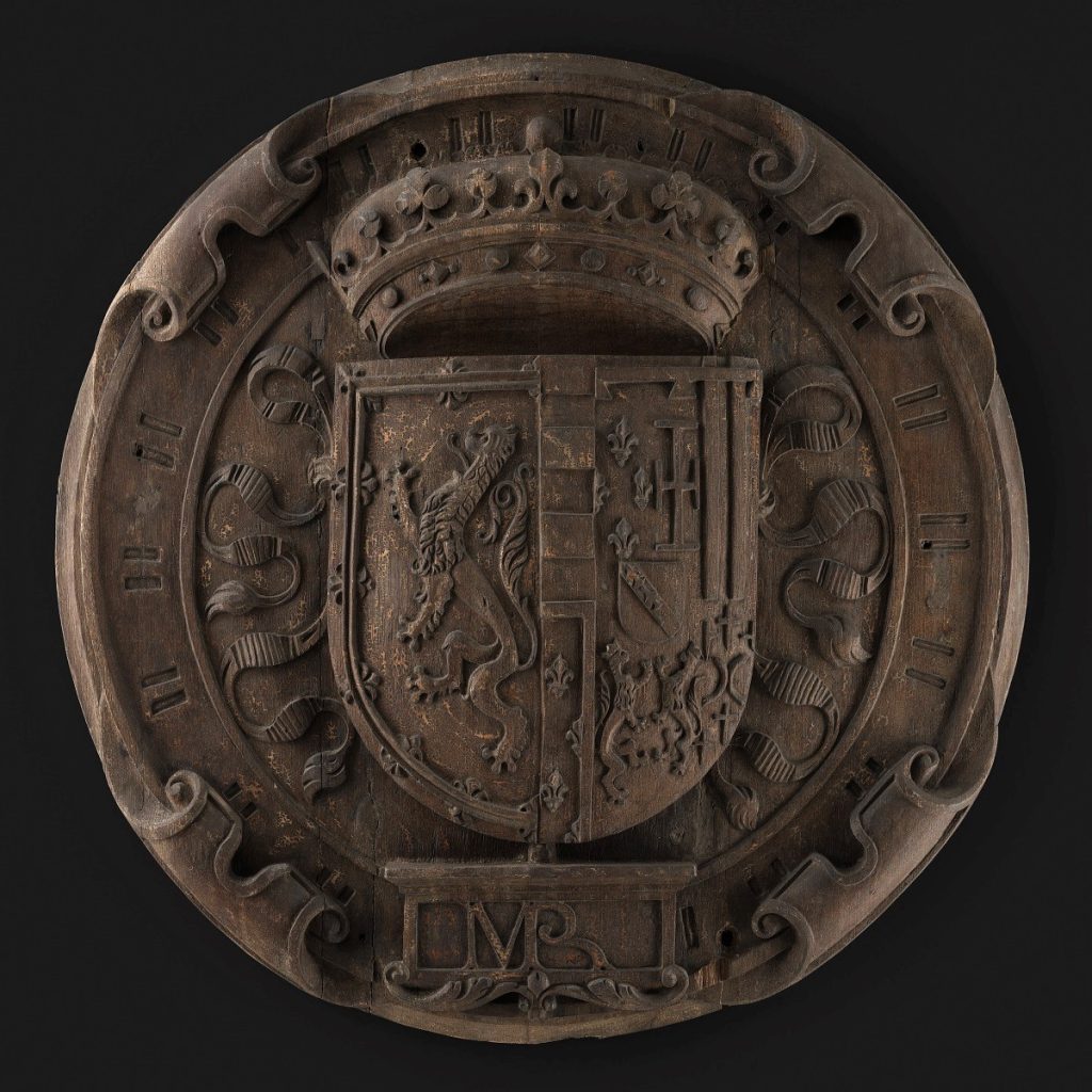 Round oak panel with boldly projecting coat of arms topped by a crown and surrounded by Latin script.