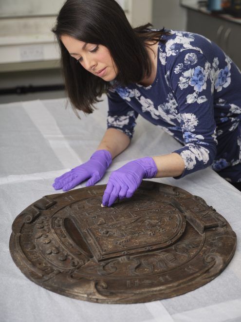 Woman with black hair in a blue floral blouse and purple gloves carefully tends to the oak panel, laid flat on a white table.