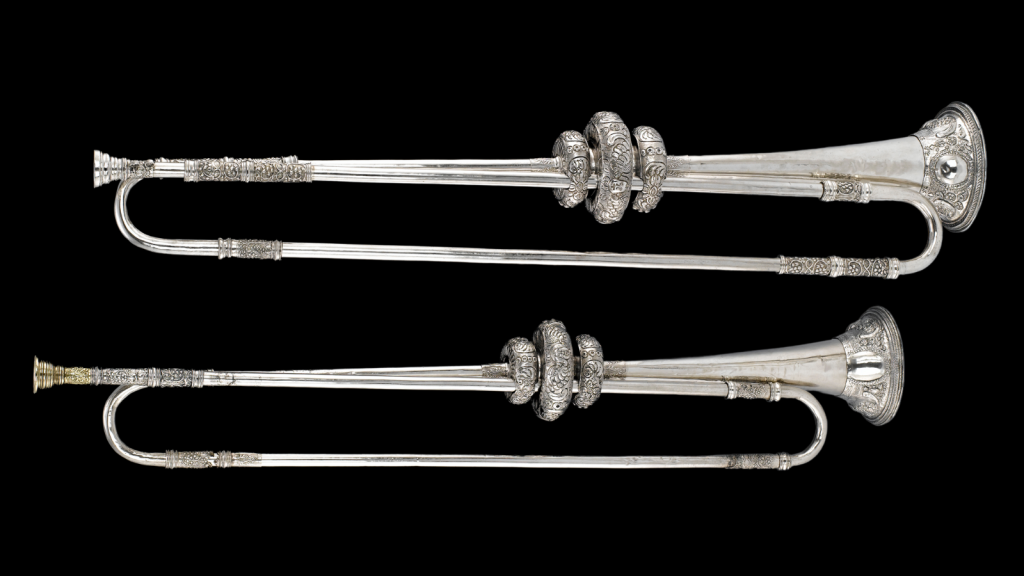 Two ornate silver trumpets, one above the other, laid out horizontally against a black background.