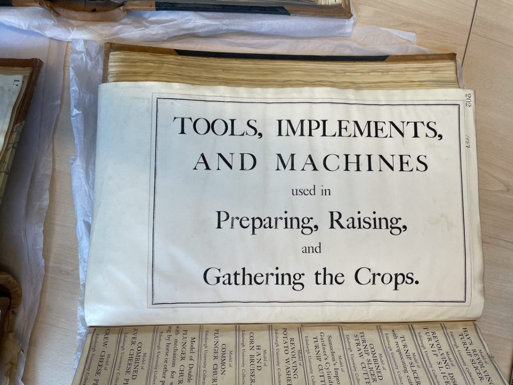Colour photo of a cream label with the description "Tools, Implements and Machines used in Preparing, Raising and the Gathering of Crops".
