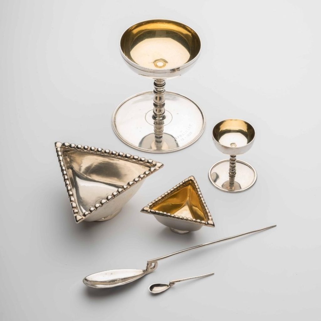 Six light silver, almost gold-tinted objects on a white surface. Two goblets, two triangular dishes, and two spoons with pin-like arms.