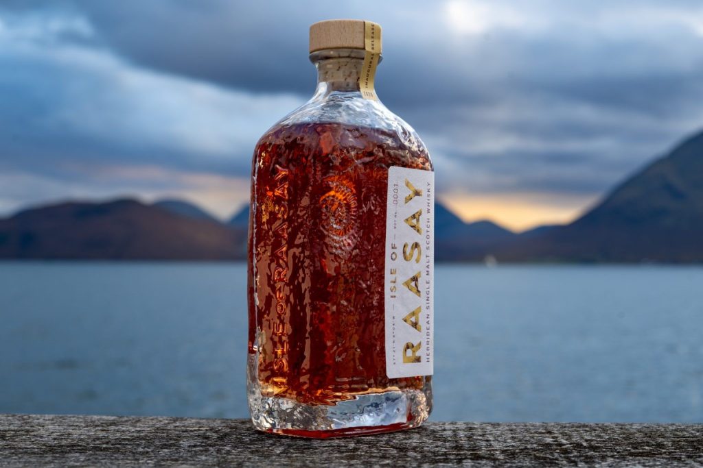 Rectangular whisky bottle with amber coloured whisky and a label reading 'Raasay' against a seaside, mountainous background.