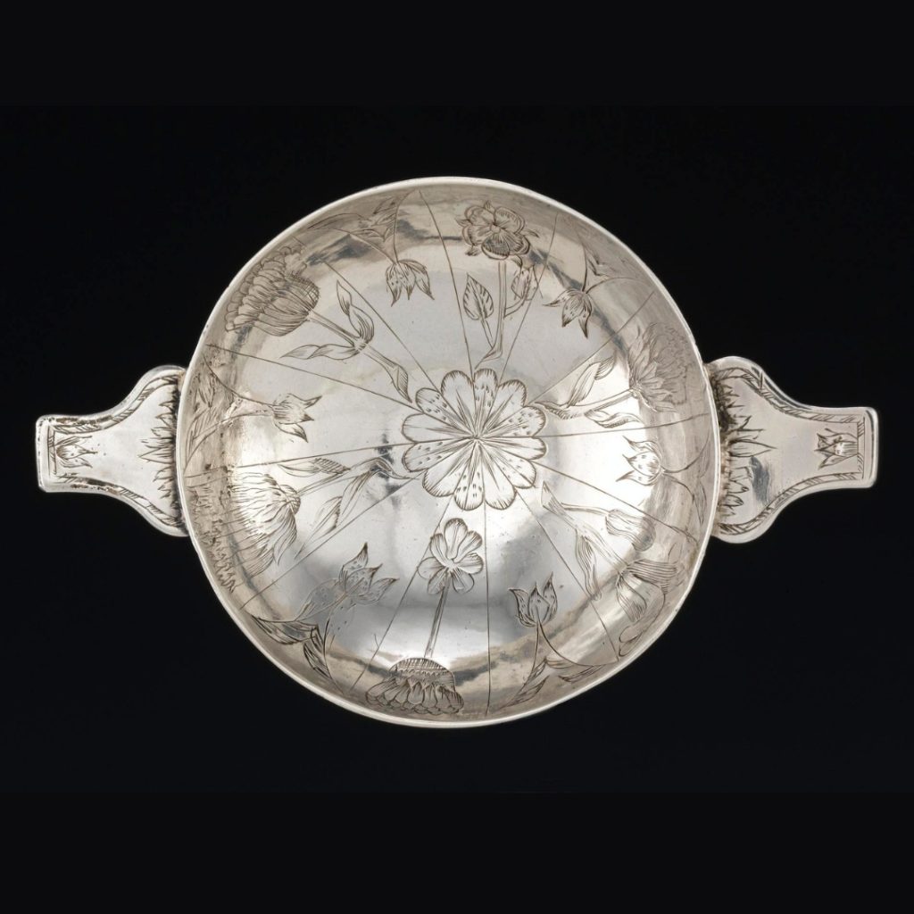 Shallow silver bowl with two solid handles viewed from above against a black background. Flower carvings decorate its inner surface.