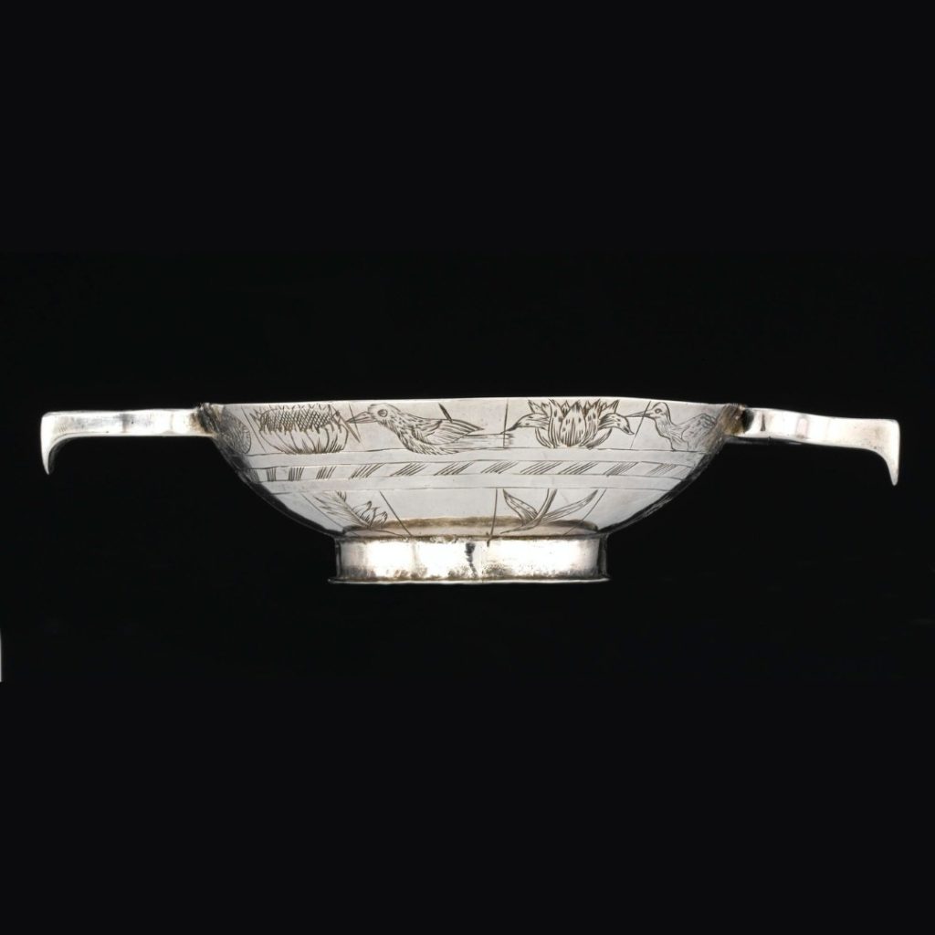 Side view of the quaich, much wider than it is deep. Carvings of birds pecking at flowers decorate its surface.