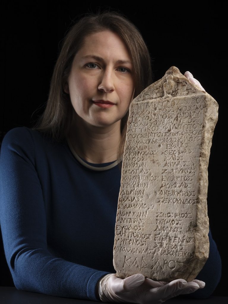Woman in a blue longsleeve shirt holds a marble slab carved with Greek letters upright against a black background.