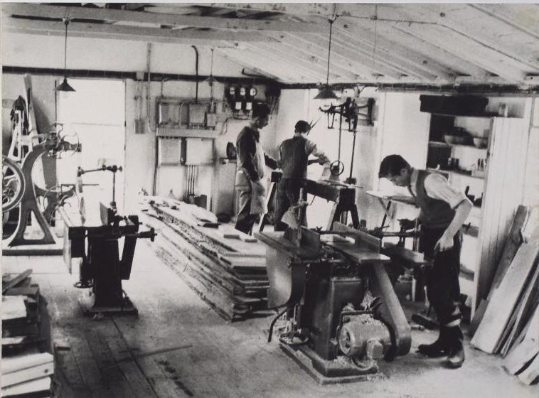 Black and white photograph of a workshop space with three men working at tables and piles of supplies on the floor.