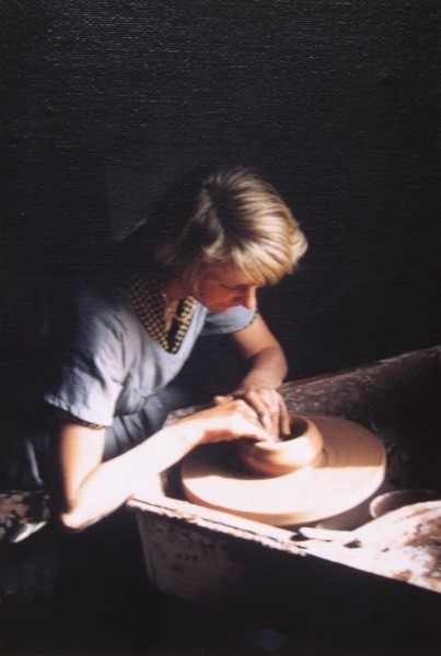 Blonde woman sits in a dark room working at a pottery wheel, her hands in the centre and looking focused.