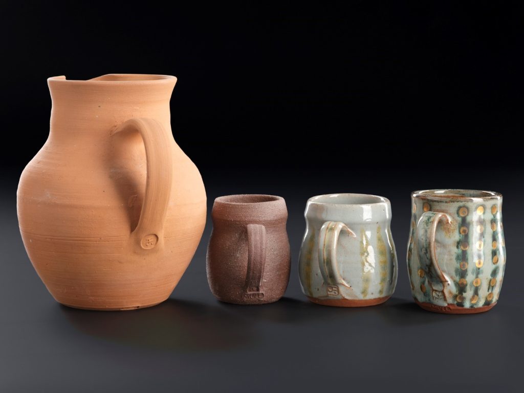 Set of four jugs in a row. On left is the largest jug, yellow-orange and looking like an ancient Greek jug. The rest look modern and the size of mugs.