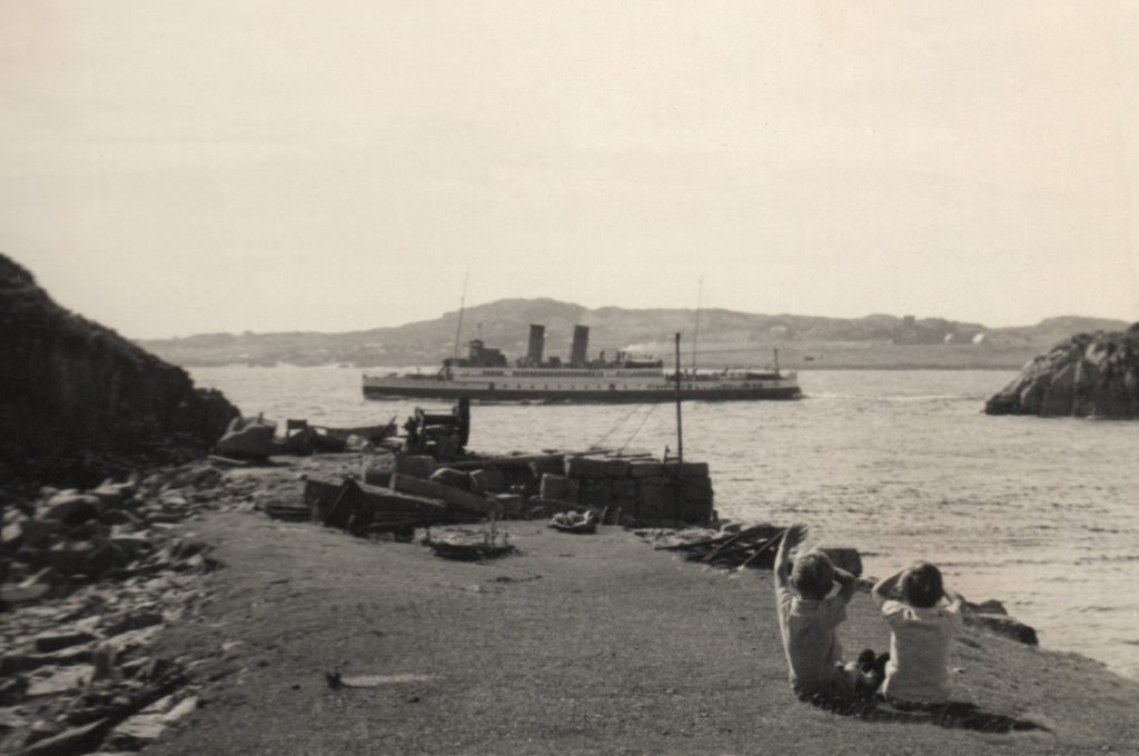 Black and white 1950s photo of a steam ship sailing through a channel being watched by children on a stone pier.